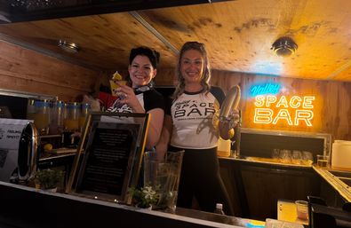 Two smiling servers behind a wood-paneled mobile bar holding a cocktail and a shaker, with a glowing neon space-themed sign and drink dispensers in the background.