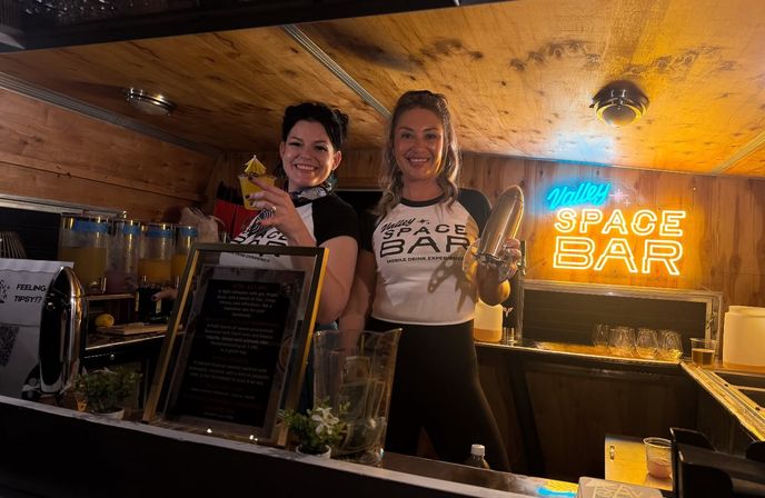 Two smiling servers behind a wood-paneled mobile bar holding a cocktail and a shaker, with a glowing neon space-themed sign and drink dispensers in the background.