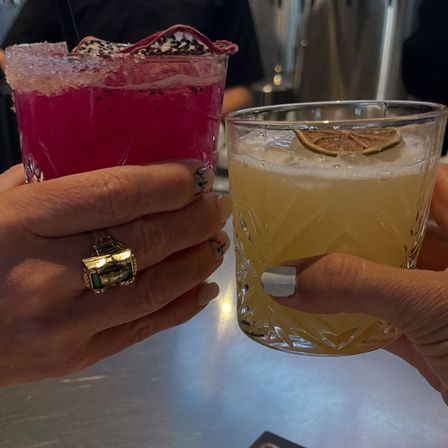 Two hands clinking cocktails over a metal bar: a bright magenta, sugar-rimmed drink with dragon fruit garnish and a pale yellow cut-glass cocktail topped with a dried lime wheel.