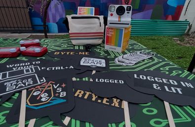 Outdoor tech-themed event table with circuit-board tablecloth, instant camera and color film box, canvas bag, sticker stack, and black speech-bubble photo props reading “LOGGED IN & LIT,” “BYTE-ME,” and “CTRL+A.”