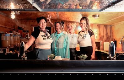 Four smiling women posing behind a cozy wooden mobile bar trailer counter with taps, drink dispensers, small potted plants and warm ambient lighting — friendly bar staff ready to serve craft drinks.