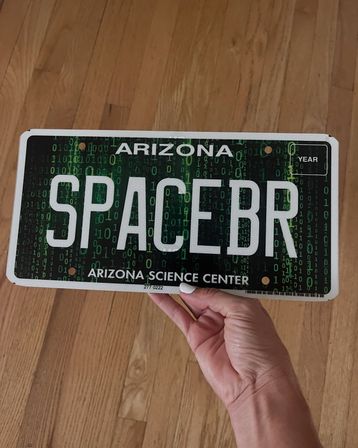Hand holding an Arizona novelty license plate reading 'SPACEBR' with a green binary-code design against a hardwood floor.