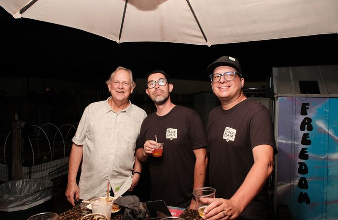 Three people smiling under a patio umbrella at night - two wearing matching black shirts and glasses hold drinks while a third in a light shirt stands beside a counter with cups and snacks; a colorful 'FREEDOM' mural is visible to the right.