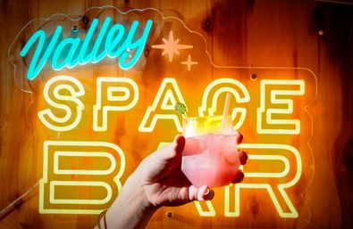Hand holding a pink, ice-filled cocktail with citrus garnish in front of a bright yellow and teal neon bar sign on a warm wooden wall.