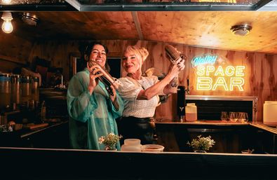 Two smiling bartenders shaking cocktail shakers behind a cozy wood-paneled mobile bar with a neon space-themed sign, drink dispensers, glassware and small herb pots on the counter.