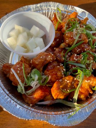 Korean-style sticky spicy fried chicken bites garnished with sesame seeds and sliced scallions, served with cubed pickled radish in a foil takeout bowl on a wooden table.