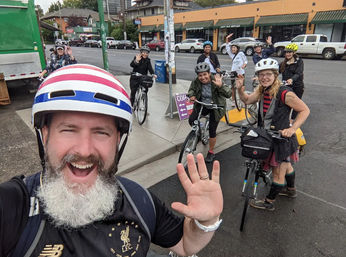 Smiling group of cyclists on a city street taking a selfie-style photo, helmets on and waving while stopped with their bikes at a curbside intersection