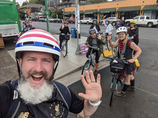 Smiling group of cyclists on a city street taking a selfie-style photo, helmets on and waving while stopped with their bikes at a curbside intersection