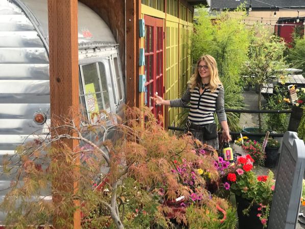 Smiling woman in a striped shirt on a plant-filled outdoor patio beside a silver vintage camper, colorful potted flowers and shrubs, wooden posts, bright yellow-red wall panels and string lights overhead.