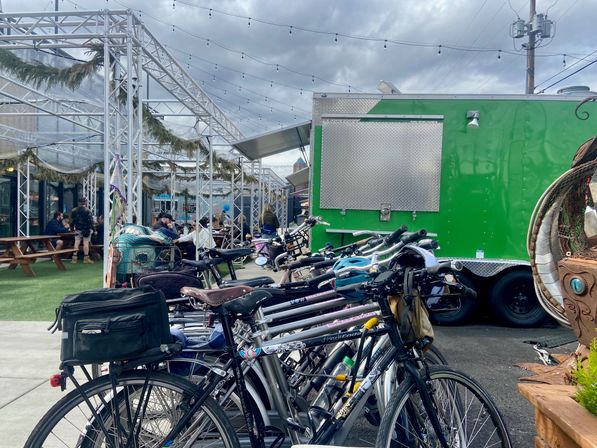 Row of parked bicycles lined up in front of a bright green food truck at a lively outdoor dining plaza with covered picnic tables, string lights and a cloudy sky