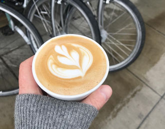 Hand in gray sweater holding a paper cup latte with leaf latte art, bike wheels and wet sidewalk in an urban outdoor setting