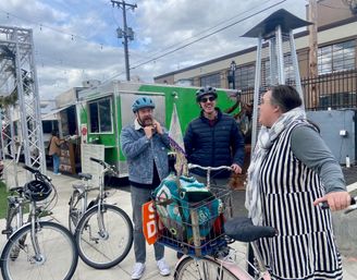 Three adults chatting beside bicycles at an urban food-truck court: two helmeted men and a woman in a striped dress standing by a bike with a basket of colorful knit items, green food truck, overhead string lights and cloudy sky.