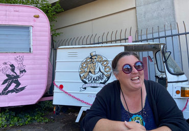 Laughing woman wearing oversized purple round sunglasses sits in front of a white vintage food truck with a pastry logo and a pink retro camper with a magician mural in an outdoor urban setting.