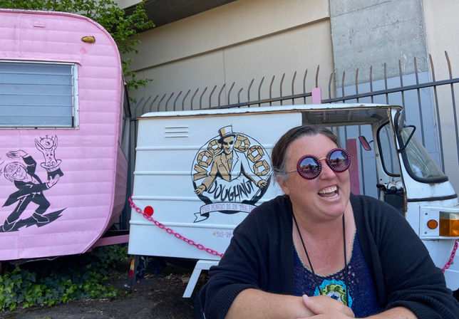 Laughing woman wearing oversized purple round sunglasses sits in front of a white vintage food truck with a pastry logo and a pink retro camper with a magician mural in an outdoor urban setting.