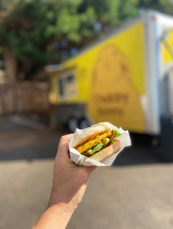 Hand holding a wrapped breakfast sandwich with egg, cheese, bacon and greens in front of a blurred yellow food truck on a sunny outdoor street