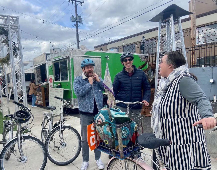 Three bicyclists chatting by a green food truck in an outdoor urban plaza; two helmeted men adjust straps while a woman in a striped dress leans on her bike with a front basket holding a colorful crocheted blanket, string lights overhead and a patio heater nearby.