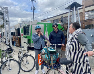 Three bicyclists chatting by a green food truck in an outdoor urban plaza; two helmeted men adjust straps while a woman in a striped dress leans on her bike with a front basket holding a colorful crocheted blanket, string lights overhead and a patio heater nearby.