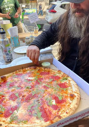 Bearded person reaching for a slice of large pepperoni and herb-topped pizza in a takeout box on a wooden outdoor sidewalk table at a casual street-side dining spot.