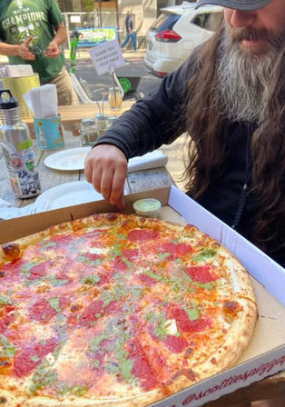 Bearded person reaching for a slice of large pepperoni and herb-topped pizza in a takeout box on a wooden outdoor sidewalk table at a casual street-side dining spot.