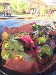 Char-grilled chicken drizzled with green herb sauce and pickled pink onions, served on brown paper with salad in a black tray on a wooden picnic table at a sunny outdoor patio.