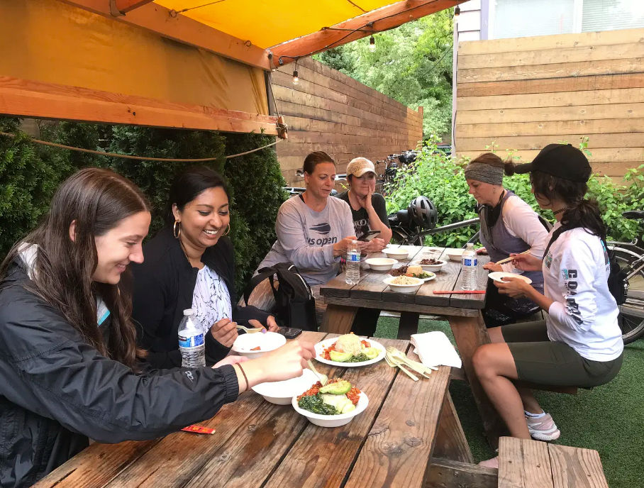 Group of friends enjoying a casual outdoor lunch at a wooden picnic table under a yellow canopy on a bike-friendly patio, sharing bowls and bottled water.