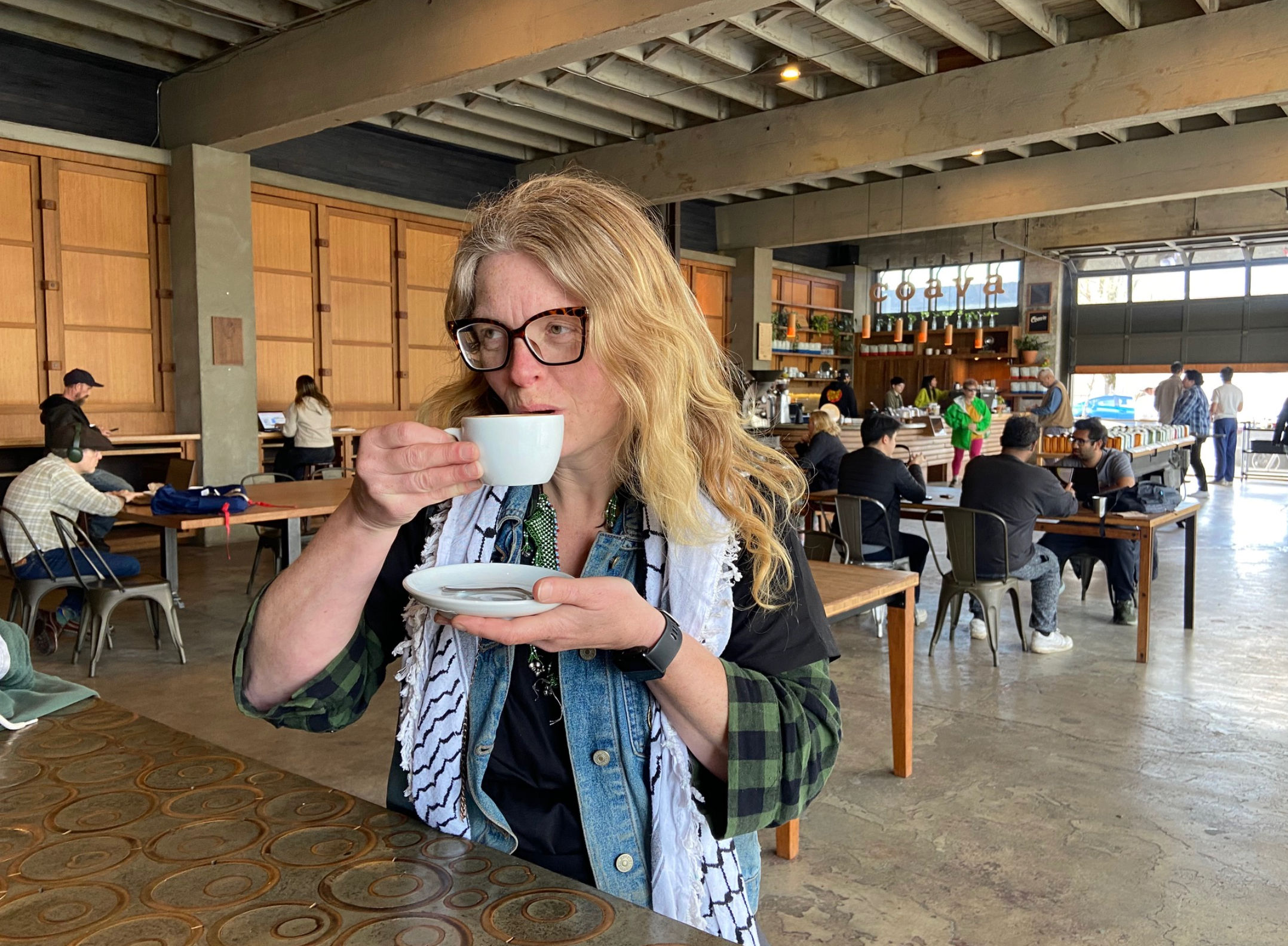Person sipping espresso from a white cup at a communal table inside an industrial-style coffee shop with exposed beams, wooden tables, baristas and customers working on laptops.
