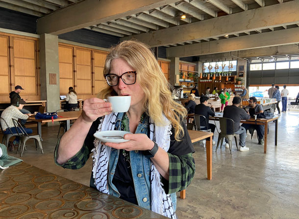 Person sipping espresso from a white cup at a communal table inside an industrial-style coffee shop with exposed beams, wooden tables, baristas and customers working on laptops.