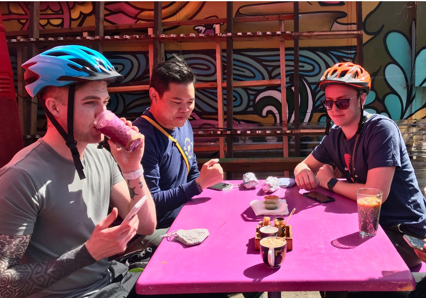 Three cyclists in helmets sit at a bright pink outdoor cafe table by a colorful mural, enjoying smoothies, coffee and small plates with foil-wrapped snacks.