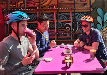 Three cyclists in helmets sit at a bright pink outdoor cafe table by a colorful mural, enjoying smoothies, coffee and small plates with foil-wrapped snacks.