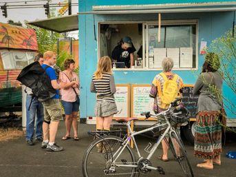 Colorful blue food truck at an outdoor market with a line of customers at the service window, chalkboard menu boards visible and a bicycle parked in front.