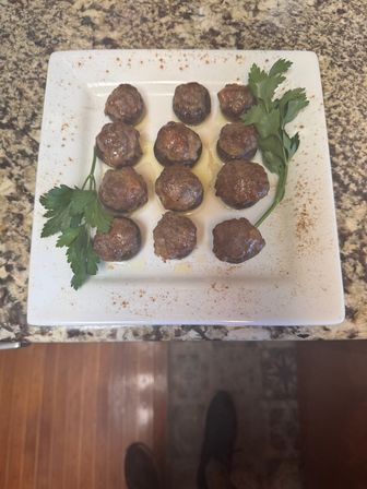 White square plate of 12 mini meatball-topped stuffed mushrooms drizzled with oil, dusted with paprika and garnished with parsley on a granite countertop
