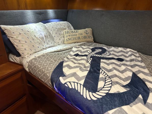 Cozy nautical-themed cabin bed with large navy anchor quilt, anchor-print sheets, 'Home is where the anchor drops' pillow, gray upholstered headboard and warm wood paneling.