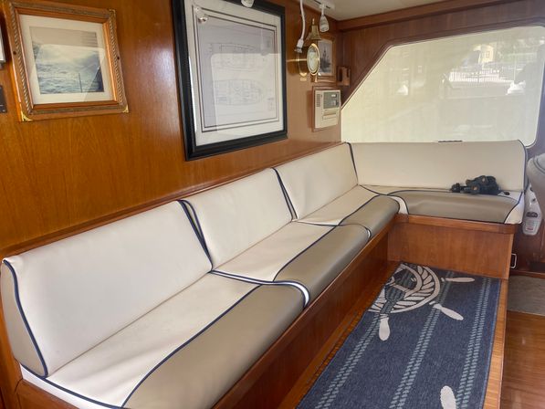 Yacht cabin lounge with L-shaped white-and-beige cushioned bench with navy piping, teak wood paneling, framed nautical prints, brass clock, ship-wheel rug and binoculars by a marina-view window