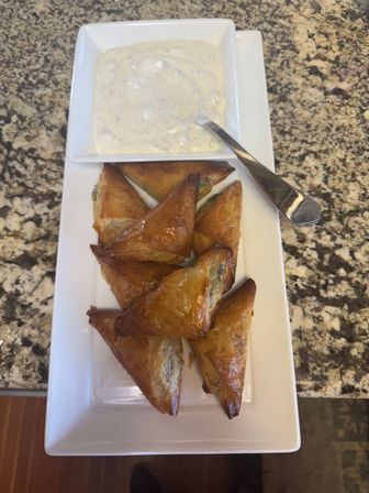 Crispy golden-brown phyllo pastry triangles (spinach-feta style) stacked on a white rectangular plate with a creamy herb yogurt dip and spoon, set on a speckled granite countertop.