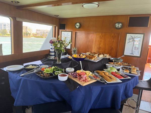 Buffet spread on a navy-clothed table inside a yacht cabin with charcuterie board, crackers, veggies, rolls, sliders, a floral centerpiece and a waterfront view
