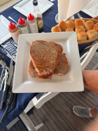 Two seared tuna steaks on a white square plate with a basket of dinner rolls, squeeze-bottle sauces and cutlery on a navy tablecloth for casual outdoor deck dining