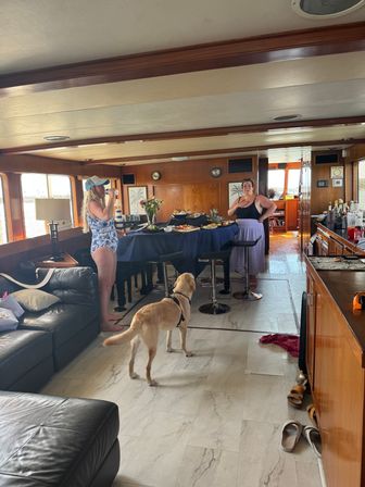 Sunlit yacht salon with wood-paneled ceiling, navy-draped buffet of snacks, two women socializing—one in a swimsuit sipping a drink—and a yellow Labrador in a harness, leather couch and water views through large windows.