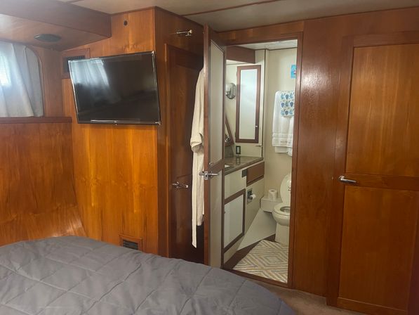 Wood-paneled yacht cabin interior with wall-mounted TV, gray quilted bed, and open door to a compact ensuite bathroom with sink, mirror, toilet, towel, and patterned bath mat.