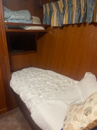 Cozy wood-paneled boat cabin bedroom with single berth, seashell-patterned quilt and pillow, wall-mounted TV beneath a shelf of folded linens, and blue-and-yellow striped curtains.