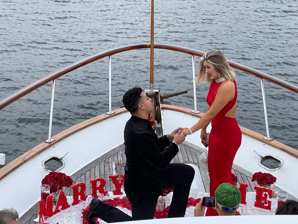 Man kneeling to propose to a woman in a red dress on a yacht deck, surrounded by red roses, 'MARRY ME' letters and a water backdrop.