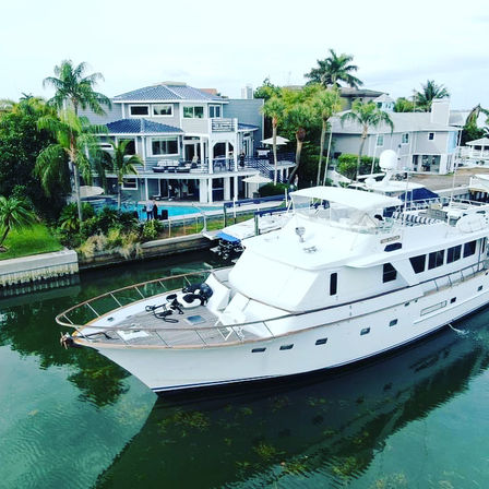 White luxury yacht gliding through a tropical waterfront canal beside a multi-level coastal mansion with palm trees, pool, and private dock