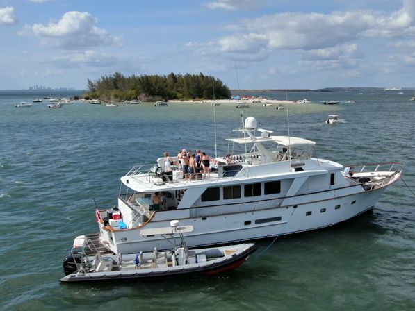 White motor yacht with a group on deck anchored near a sandy island and scattered boats in a sunny coastal bay