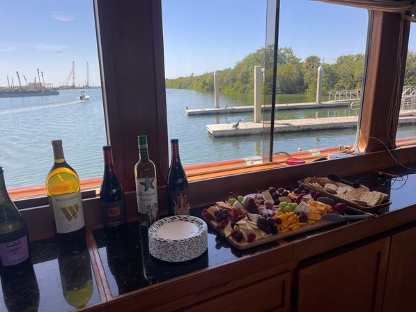 Waterfront charcuterie and wine spread on a boat counter — bottles of wine beside a wooden board of cheeses, cured meats, grapes and crackers, framed by windows overlooking a marina dock, calm water and tree-lined shore under a clear blue sky.