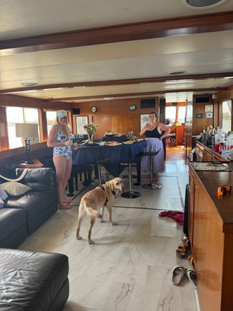Sunlit wood-paneled yacht salon with a buffet-covered table, two women in summer attire near panoramic windows and a yellow Labrador standing on light tile flooring.