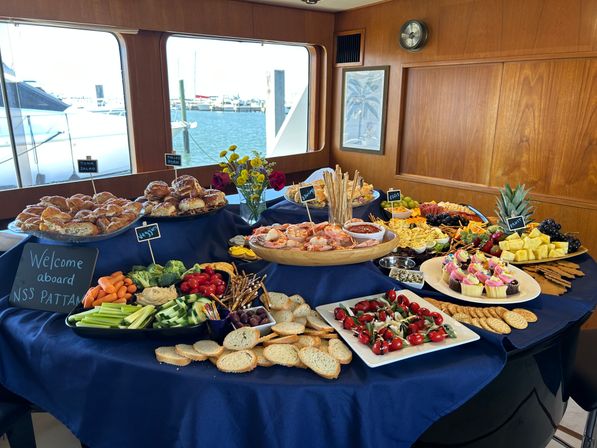 Yacht brunch buffet at a marina with a navy-draped table of shrimp cocktail, croissants, vegetable and fruit platters, caprese skewers, crackers, cupcakes and dips, seen against windows overlooking docked boats and water.
