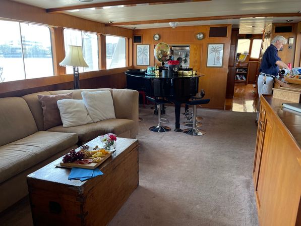 Wood-paneled luxury yacht salon with a beige sectional, trunk coffee table holding a charcuterie board and flowers, glossy black grand piano doubled as a bar with chrome stools, large windows with a waterfront view, and a person at the galley preparing drinks.