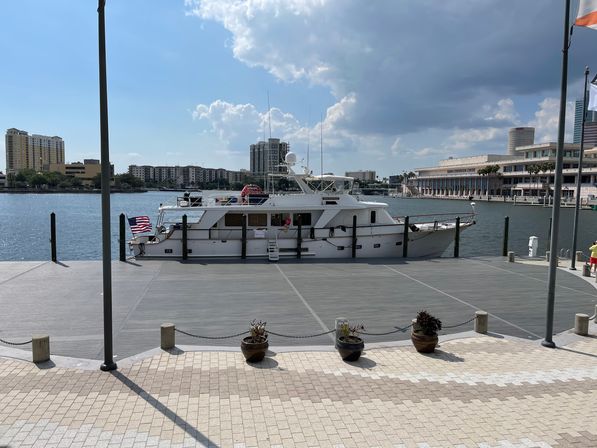 White motor yacht docked at a sunny downtown waterfront marina, American flag on the stern, paved promenade with planters in the foreground and urban skyline and convention-center style building across the bay.