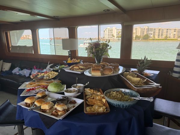 Yacht buffet spread with sliders, sandwiches, chips, dips, cheese and fruit on a navy-clothed table, vase of flowers centerpiece and waterfront condos visible through large windows.