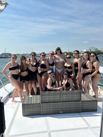 Group of friends in bikinis posing on a yacht deck during a sunny summer boat party, marina and sailboats visible on the harbor skyline