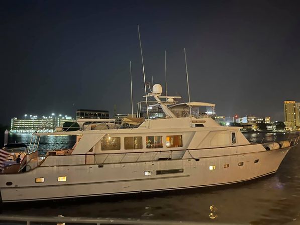 White luxury motor yacht docked at a city waterfront marina at night, warm cabin lights reflecting on calm harbor water with illuminated buildings and parking structure in the background.